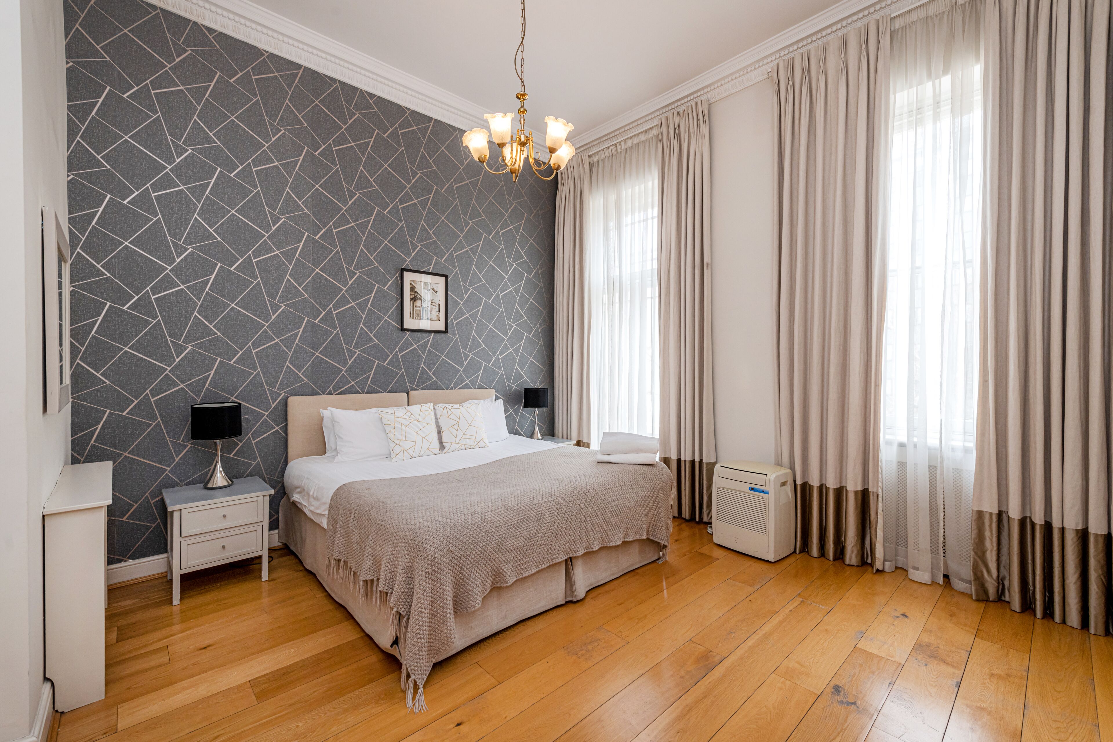 A bedroom with a modern design, featuring a bed with a beige bedspread, a nightstand with a lamp, a wall-mounted picture, and a large window with curtains.