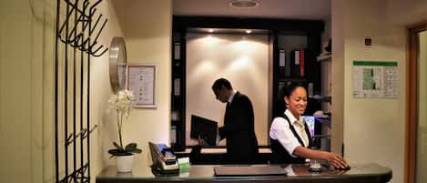 A reception desk with a plant, a phone, and a laptop. There is a fire extinguisher and a sign on the wall.