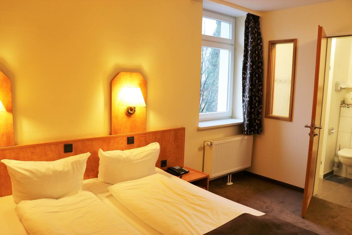 Een hotelkamer met een bed, bedlampen, een raam met gordijnen, een radiator en een deur naar een badkamer.