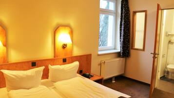 Habitación de hotel con cama, lámparas de noche, ventana con cortinas, radiador y puerta que da al baño.