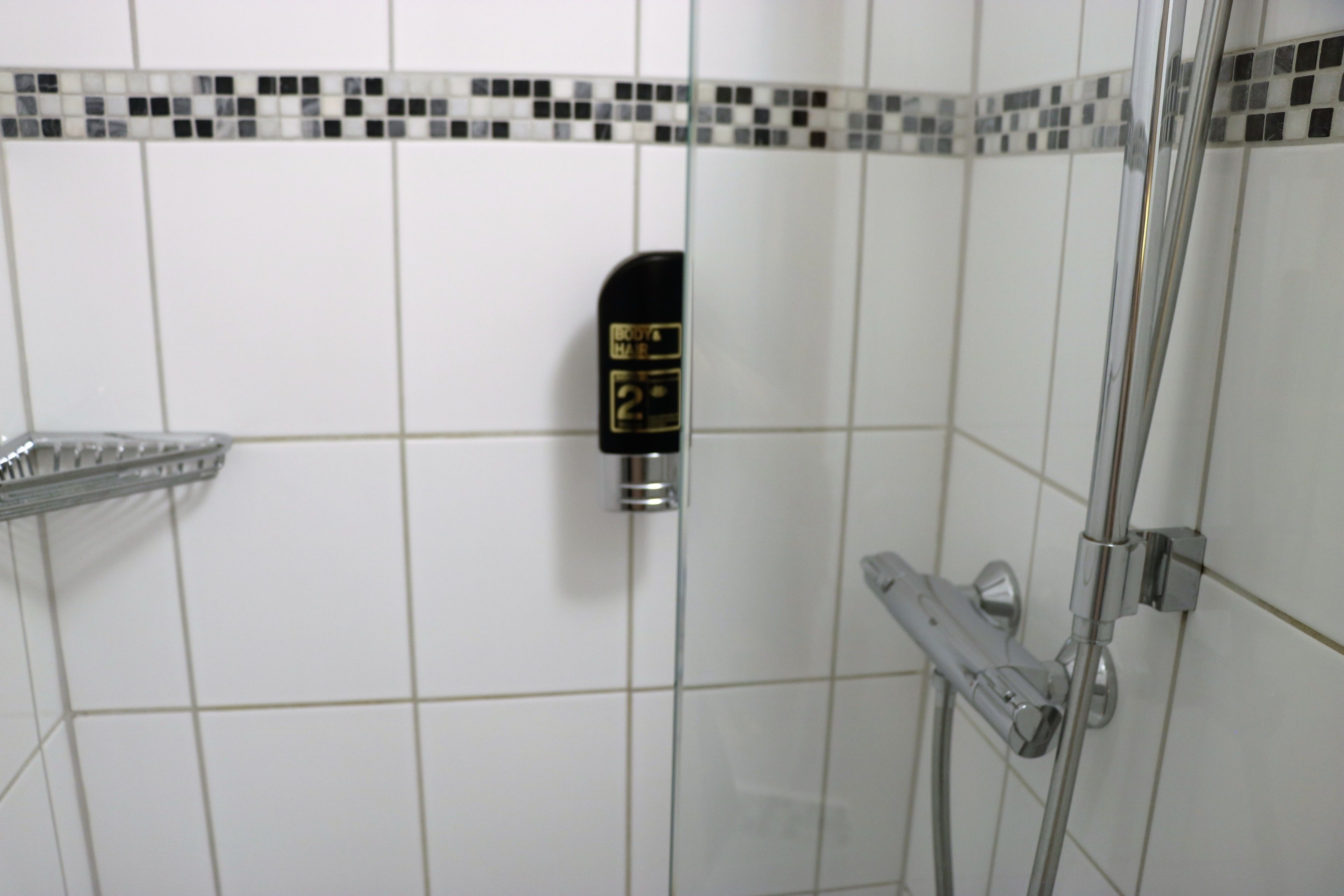 Zona de ducha con cabezal de ducha manual montado en la pared, mampara de vidrio y suelo de baldosas.