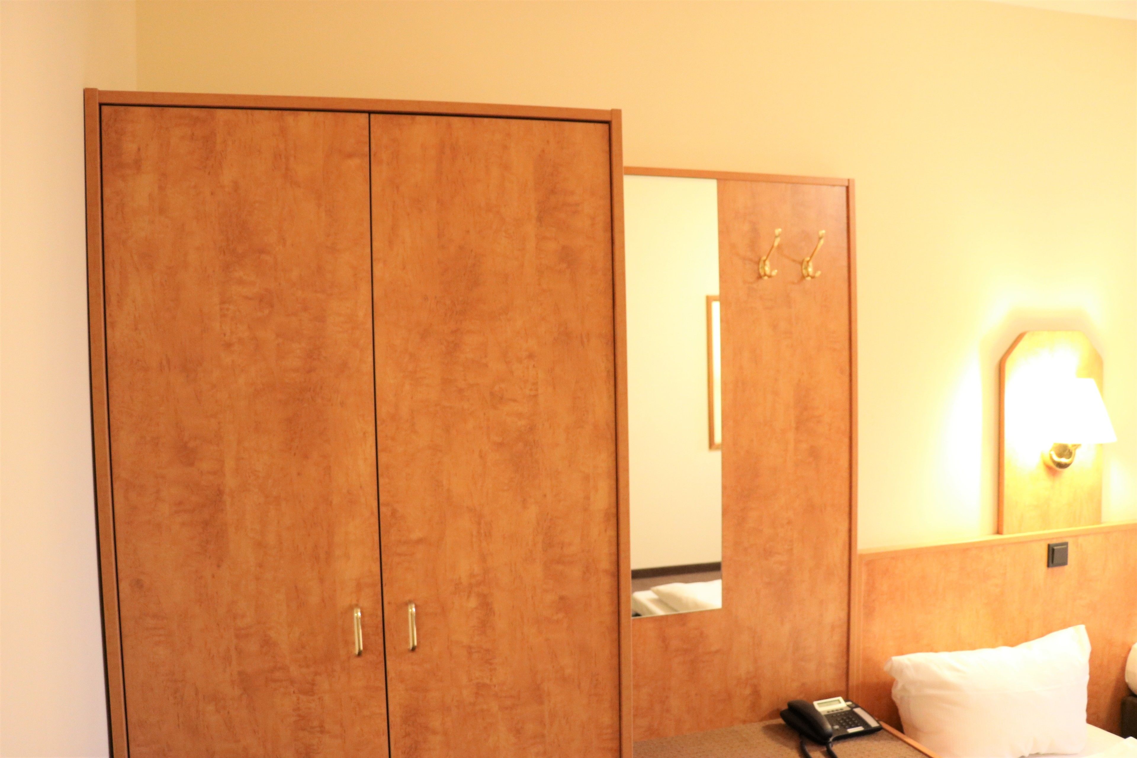 Una camera d'albergo con un armadio in legno, un comodino con una lampada e uno specchio che riflette parte della stanza.