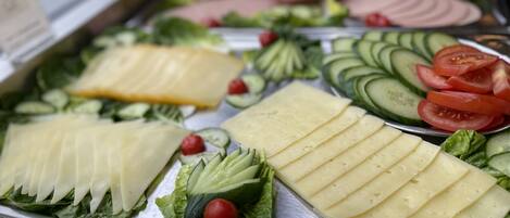 Un bufé con diversos alimentos, incluyendo queso en rodajas, pepinos, tomates y hojas verdes.