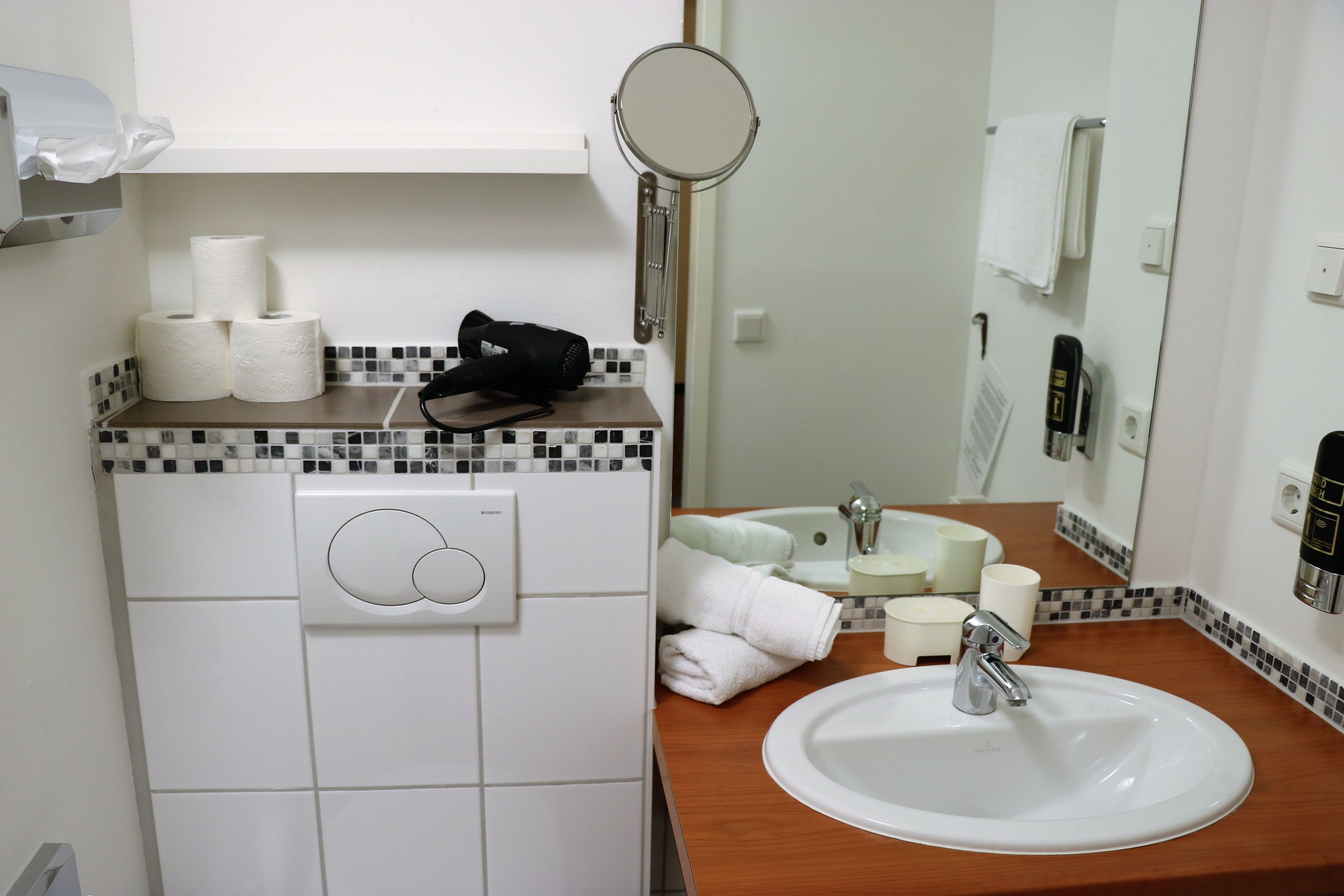 Un baño con lavamanos blanco, espejo, toallero y dispensador de papel higiénico.