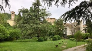 A lush garden with a pathway leading to a building with a clock tower. - B&B Villa al Parco (Sala Baganza)