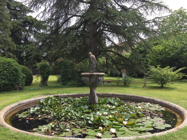 A circular pond with lily pads and a statue in the center, surrounded by lush greenery and trees. - B&B Villa al Parco (Sala Baganza)