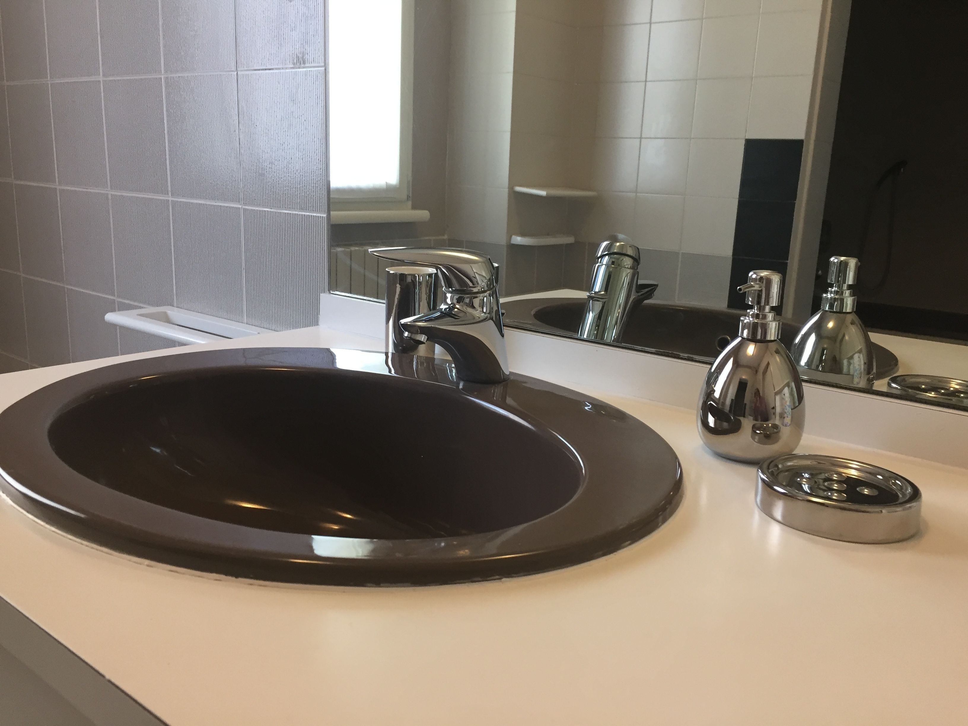 Ein modernes Badezimmer mit einem runden, schwarzen Waschbecken, chromfarbenen Wasserhähnen und einem Spiegel, der Waschbecken und eine Seifenflasche reflektiert.