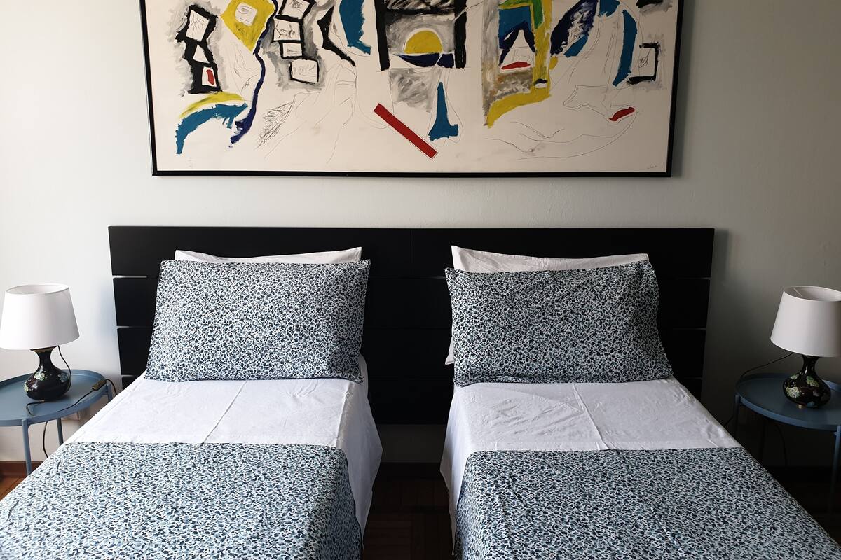 A hotel room with two beds, white bedding, a black headboard, and a large abstract painting on the wall.