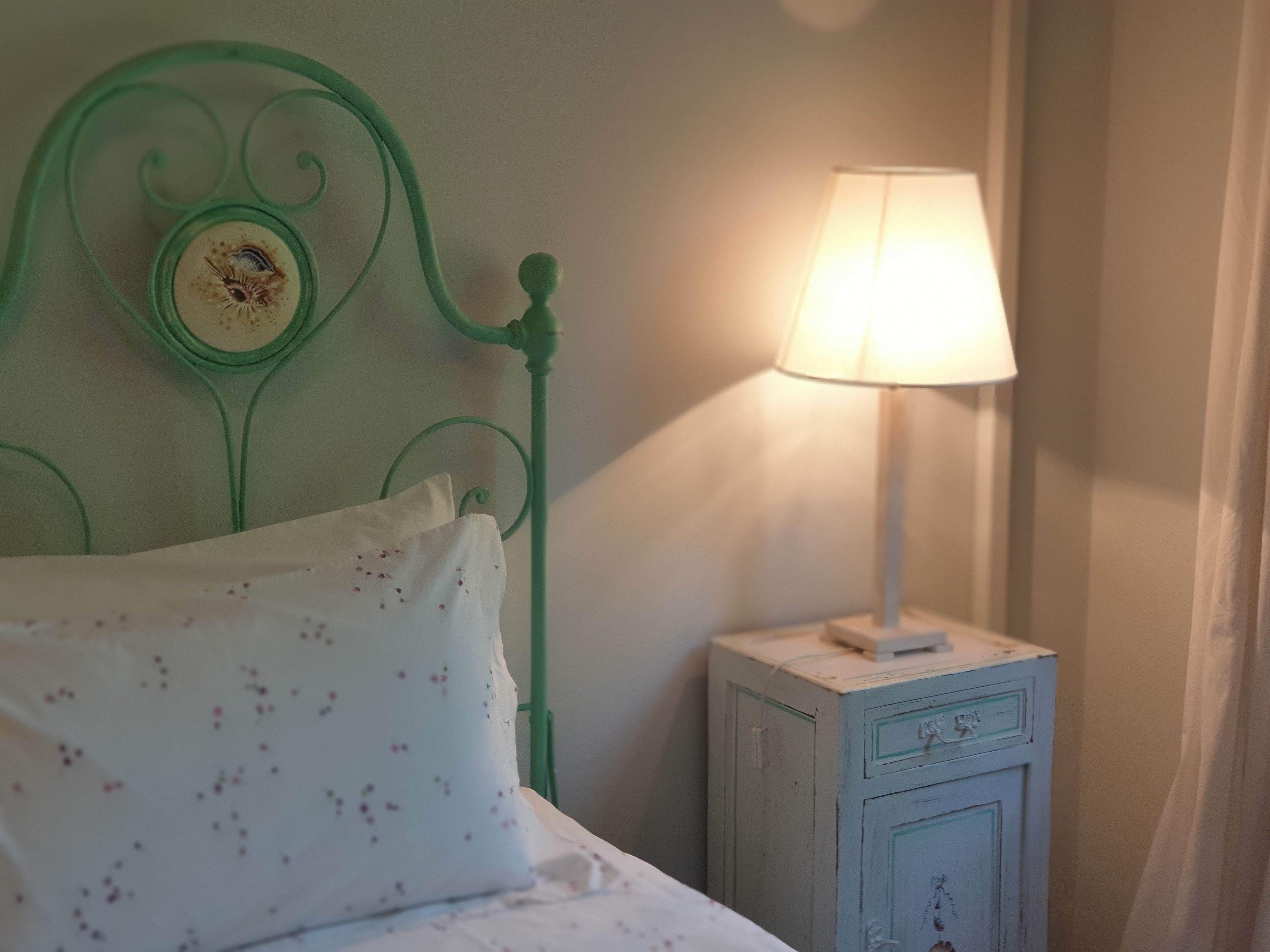 Uma cama com cabeceira de metal verde, um travesseiro branco com padrão floral e uma mesa de cabeceira com uma luminária.