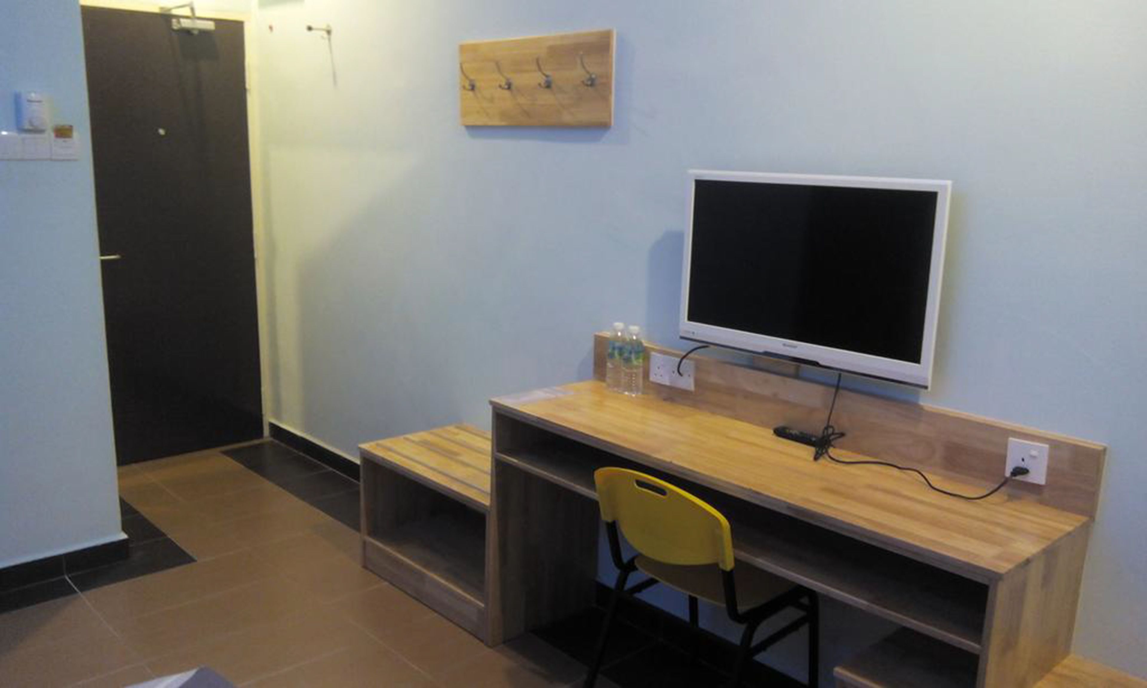 Una stanza piccola e modesta con una scrivania in legno, una sedia gialla, una televisione e un gancio per cappotti a parete.