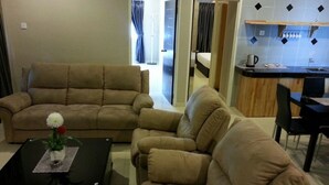A living room with two beige sofas, a coffee table, and a small table with a flower vase. There is a kitchen area with a sink and cabinets visible in the background. - Hotel Suria 18 (Ipoh)