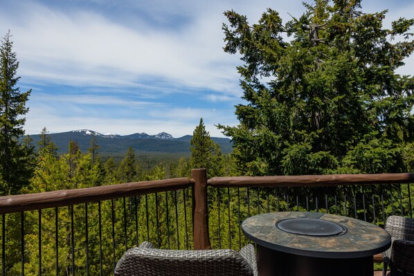 A deck with a round table and wicker chairs, overlooking a forest and mountains.