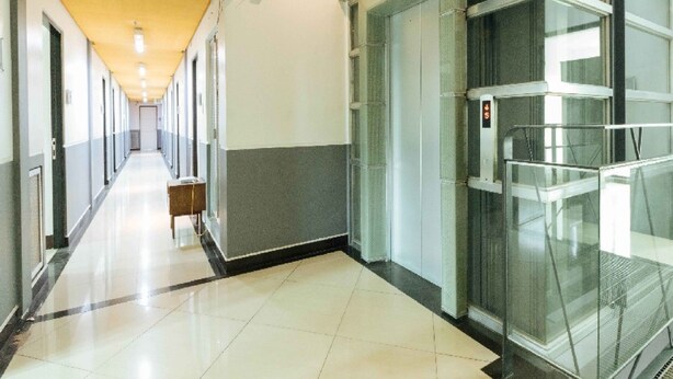 Um corredor moderno de hotel com elevador de vidro, um banco e um longo corredor com portas.