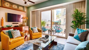 Ein Wohnzimmer mit einer Couch, Sesseln, einem Couchtisch, einem Fernseher und einem Balkon mit Sitzgelegenheiten.