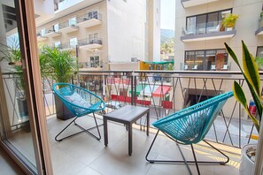 En balkon med to blå vævede stole og et lille bord, med udsigt over en by silhuet med bygninger og en butik med rødt tag.