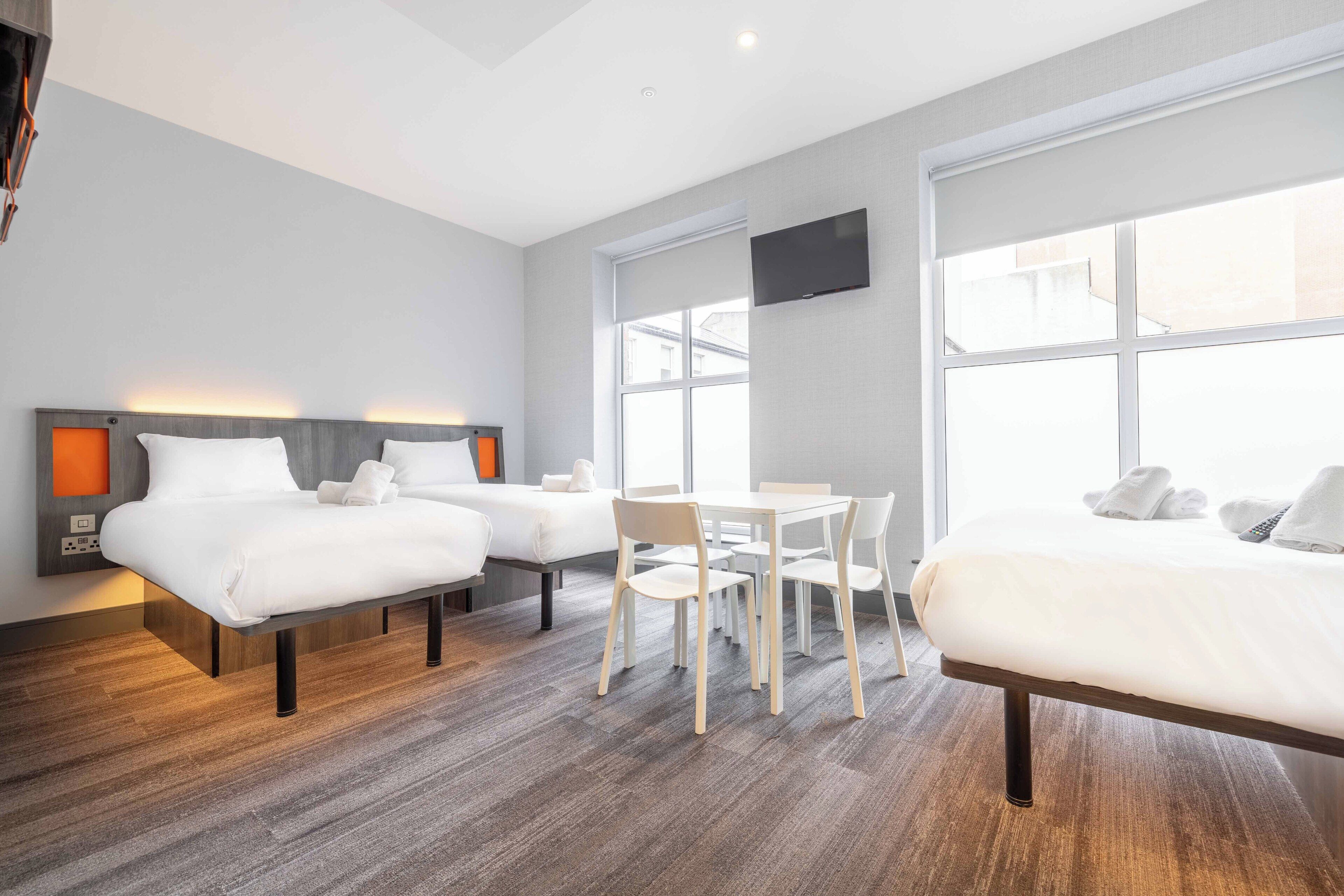 A hotel room with two beds, a dining table, chairs, and a TV mounted on the wall.