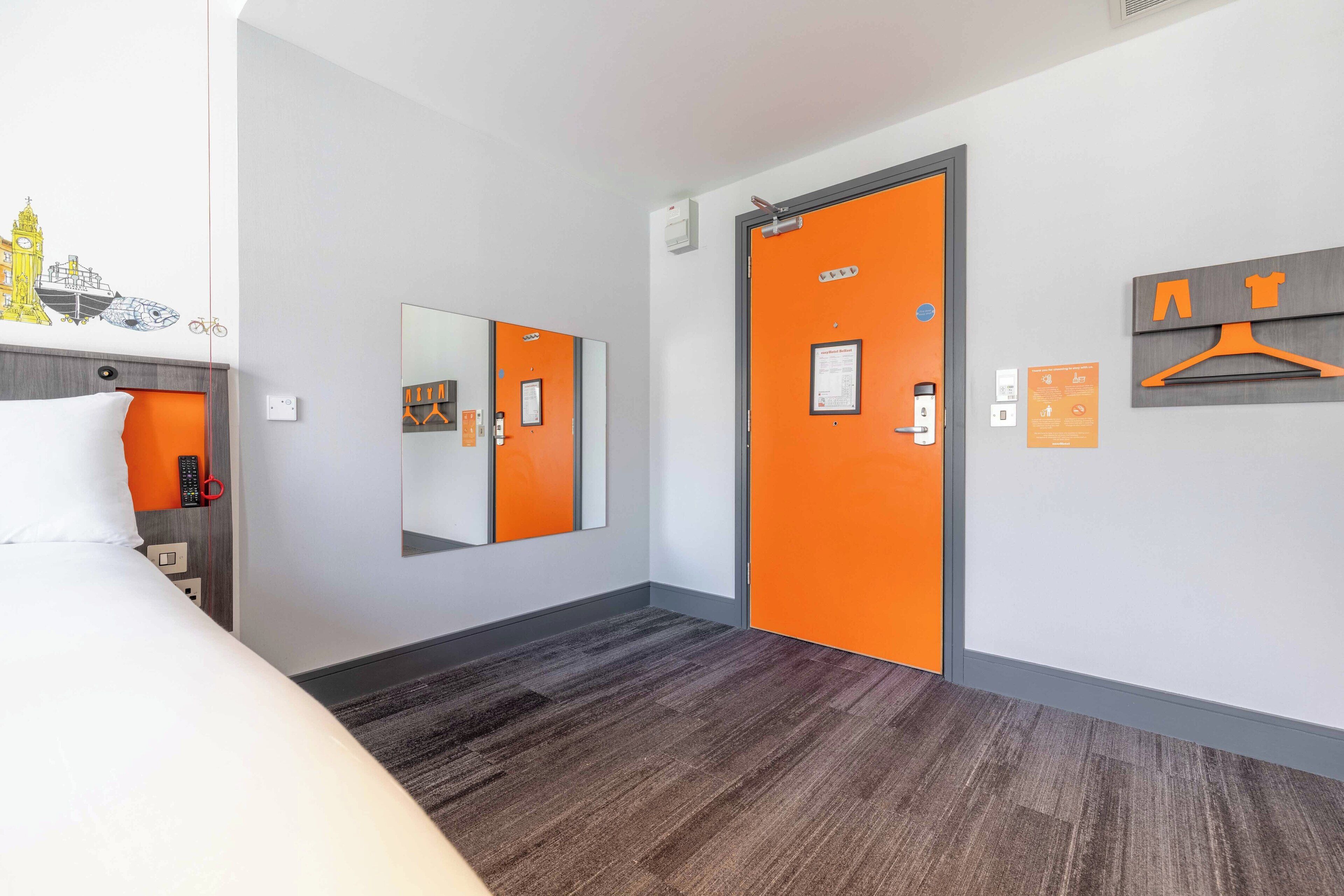 Une chambre d’hôtel avec un lit, une porte orange, un miroir et un porte-manteau fixé au mur.