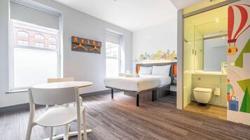 Habitación de hotel moderna con una cama, una mesita, una silla, un ventanal con vistas a edificios y un baño con inodoro y espejo.