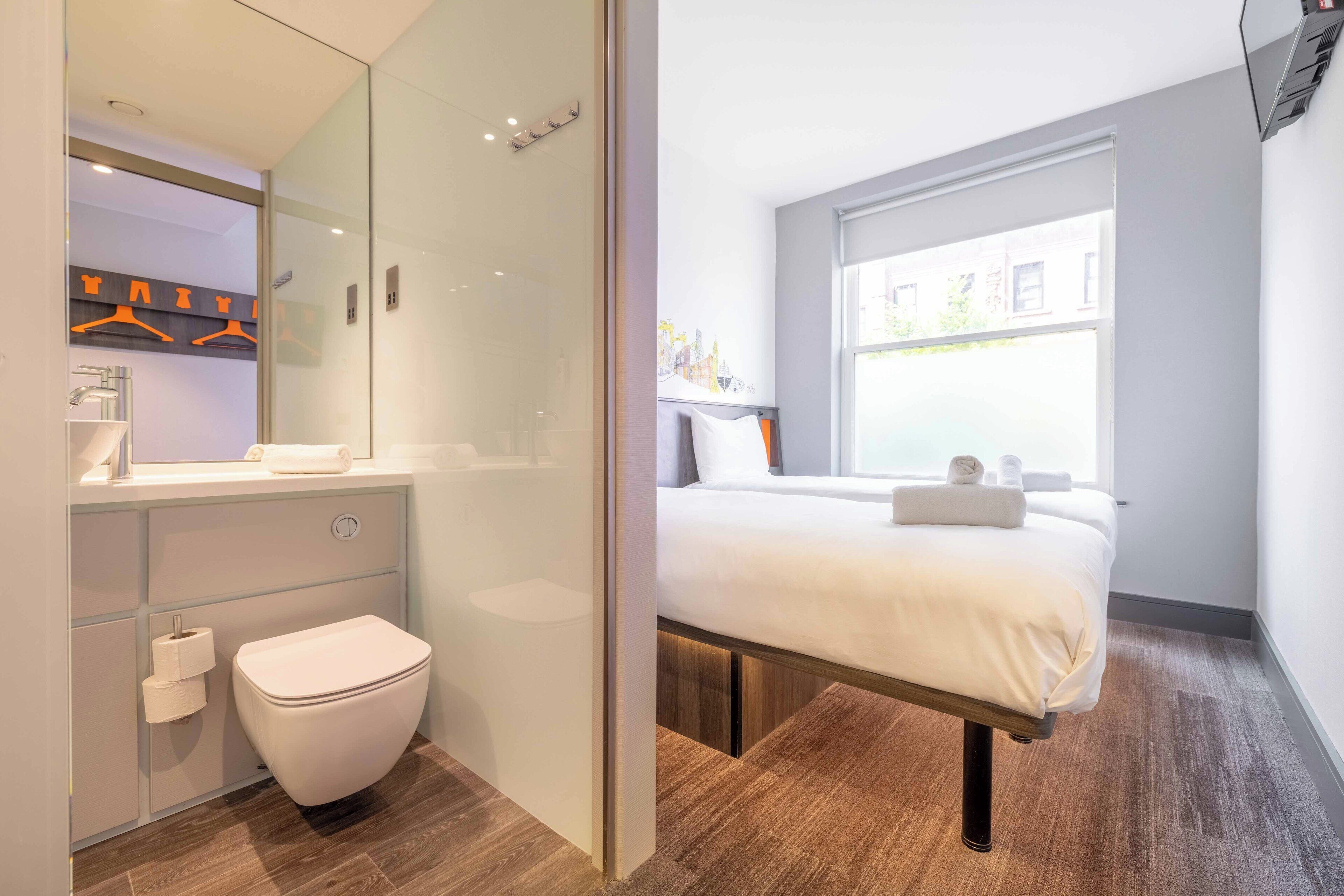 Una habitación de hotel moderna con un ventanal grande, una cama, zona de ducha y baño con inodoro y lavabo.