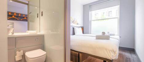 Una habitación de hotel moderna con un ventanal grande, una cama, zona de ducha y baño con inodoro y lavabo.
