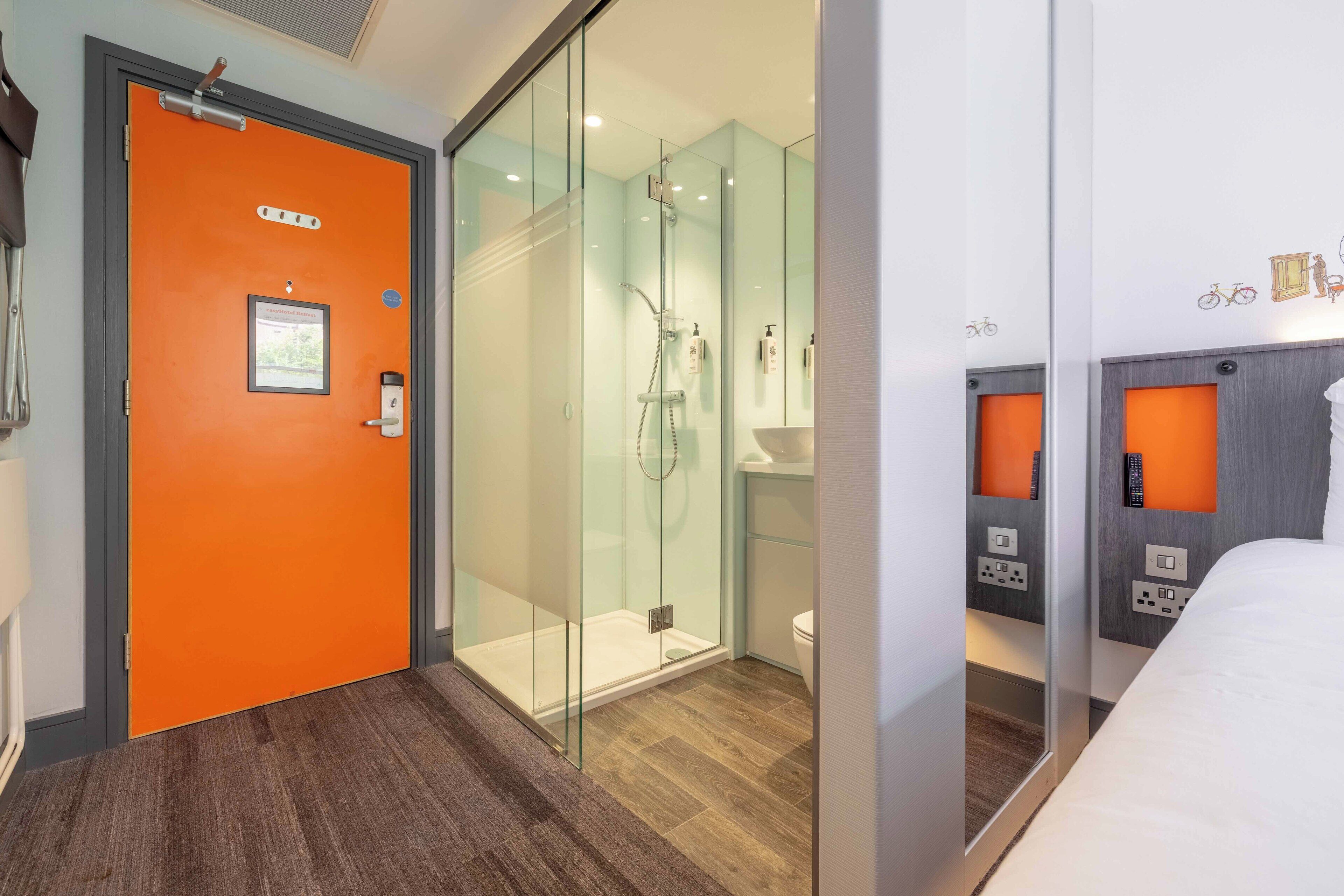 Ein Hotelzimmer mit einer orangefarbenen Tür, einer Duschkabine aus Glas, einem Holzboden, einem weißen Bett und einem Spiegel, der den Raum reflektiert.