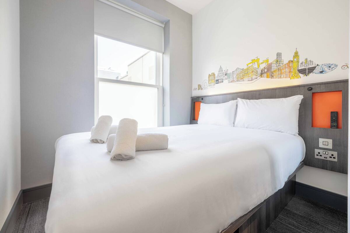 A hotel room with a large bed, white linens, two rolled towels, a cityscape mural on the wall, a window, and a nightstand with a lamp and remote control.