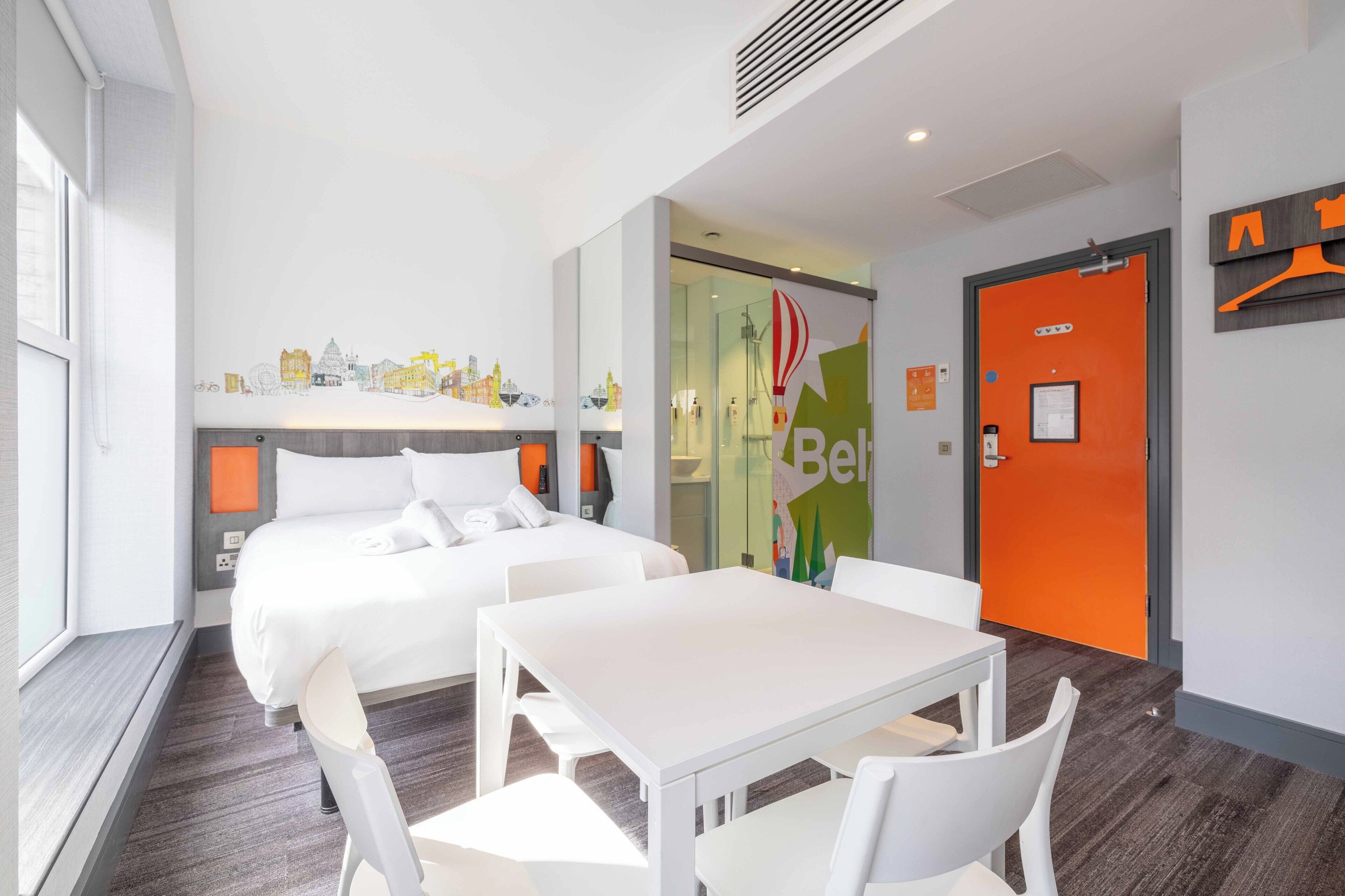 A modern hotel room with a large bed, a small table, and a seating area. There is a glass door leading to a bathroom, and a bright orange wall with a hanging coat rack.