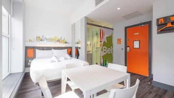 Ein modernes Hotelzimmer mit einem großen Bett, einem kleinen Tisch und einer Sitzecke. Eine Glastür führt ins Badezimmer, und eine leuchtend orangefarbene Wand ziert einen hängenden Kleiderhaken.