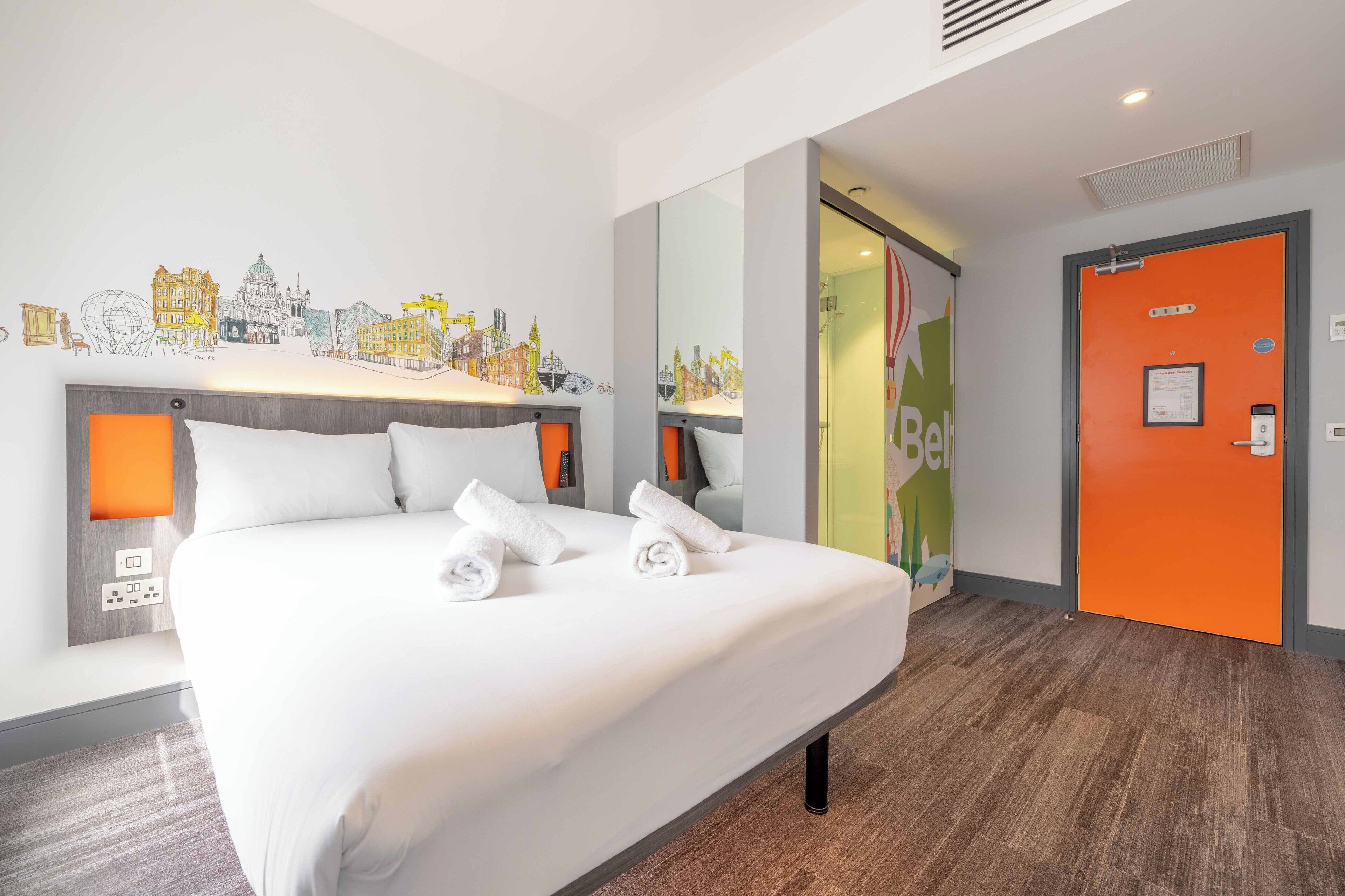 A hotel room with a large bed, two bedside tables with lamps, a cityscape mural on the wall, and an orange door.
