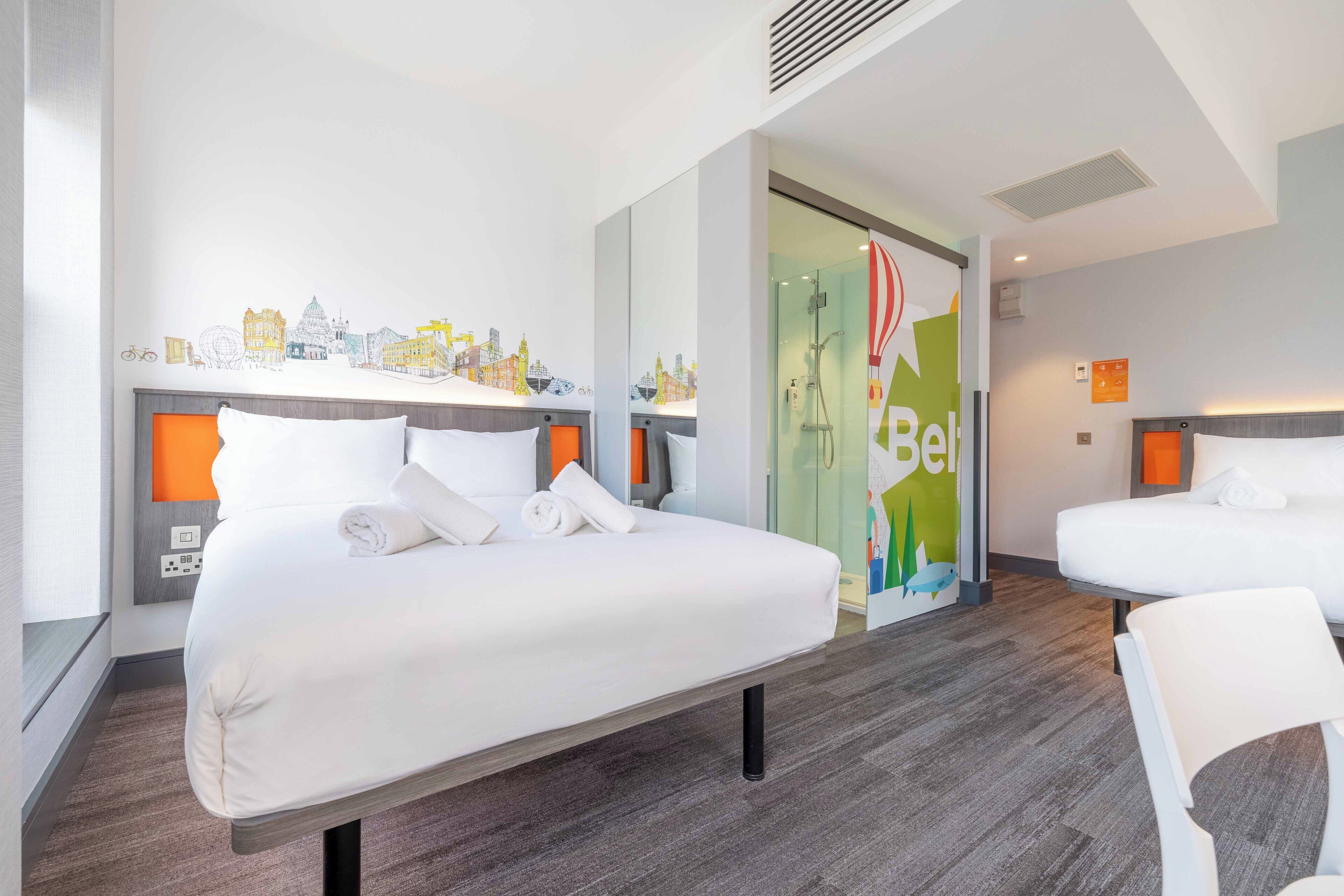 A hotel room with two beds, a shower area, and a wall with a cityscape mural.