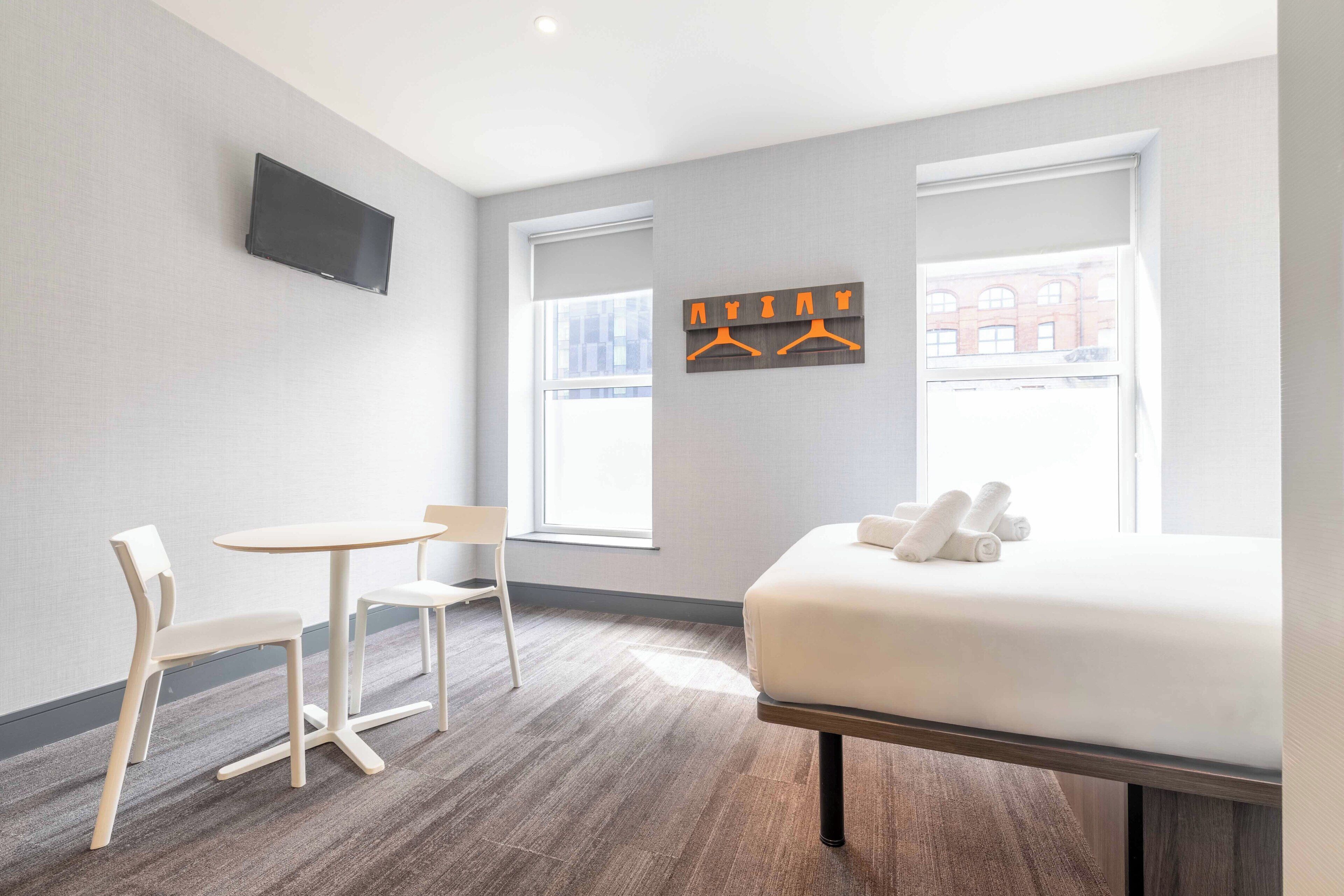 Et hotelværelse med en seng, et lille bord, to stole, et tv ophængt på væggen og et vindue med udsigt til bygninger.