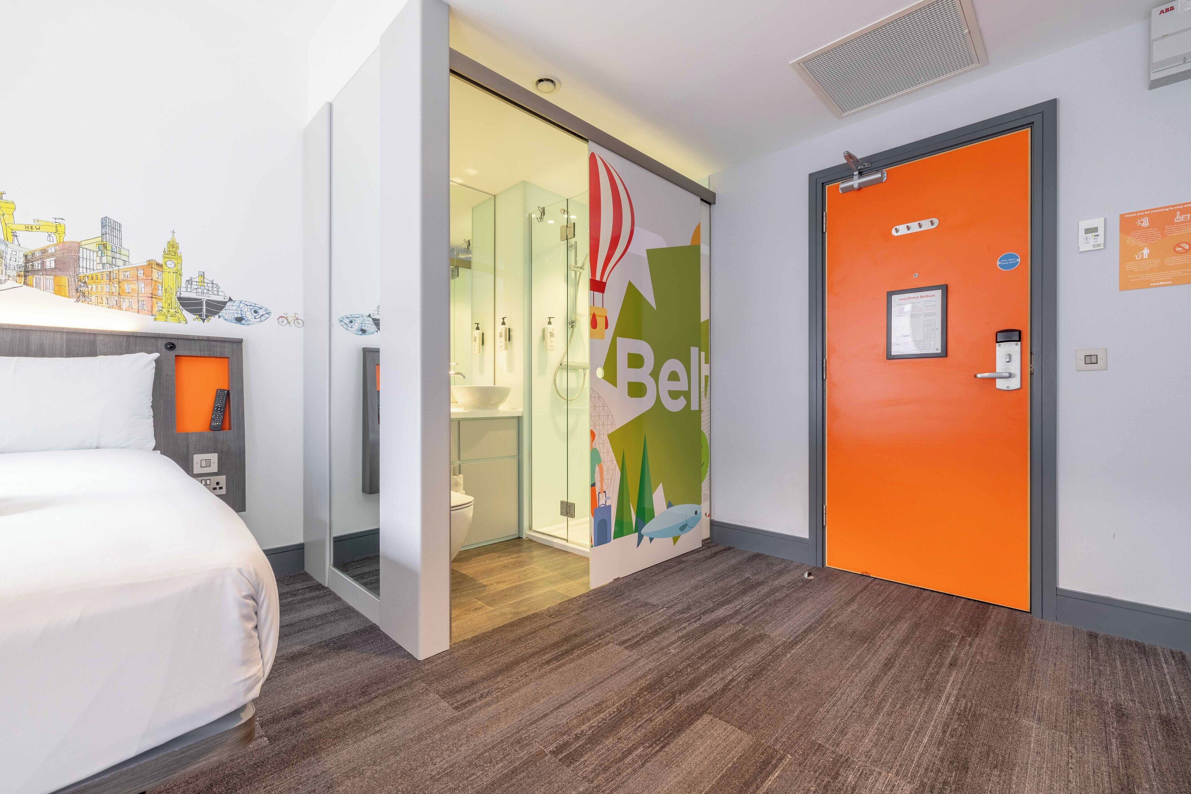Una habitación de hotel con cama, un baño con cabina de ducha de vidrio y una puerta naranja.