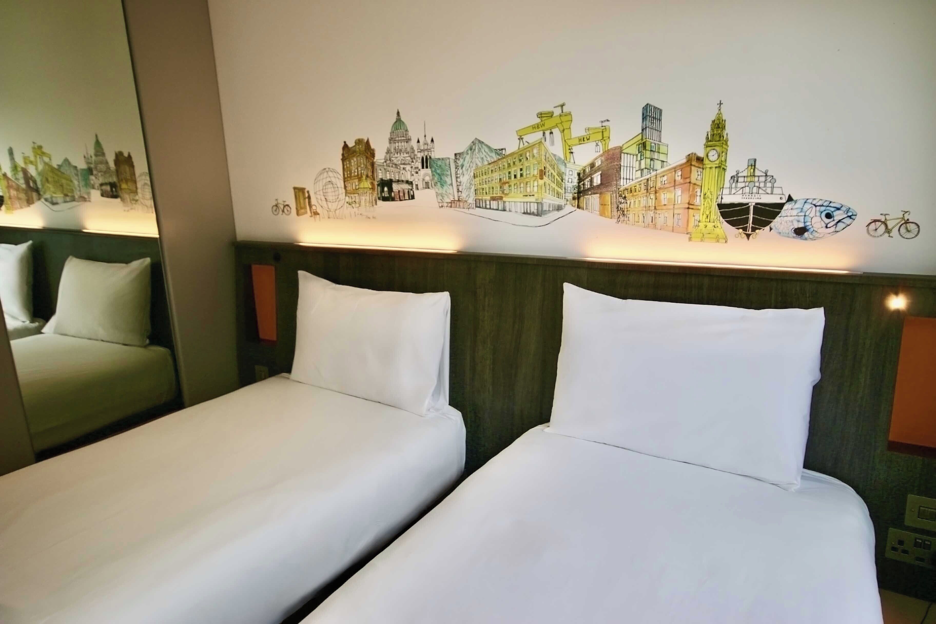 A hotel room with two beds, a cityscape mural on the wall, and a mirror reflecting the room.