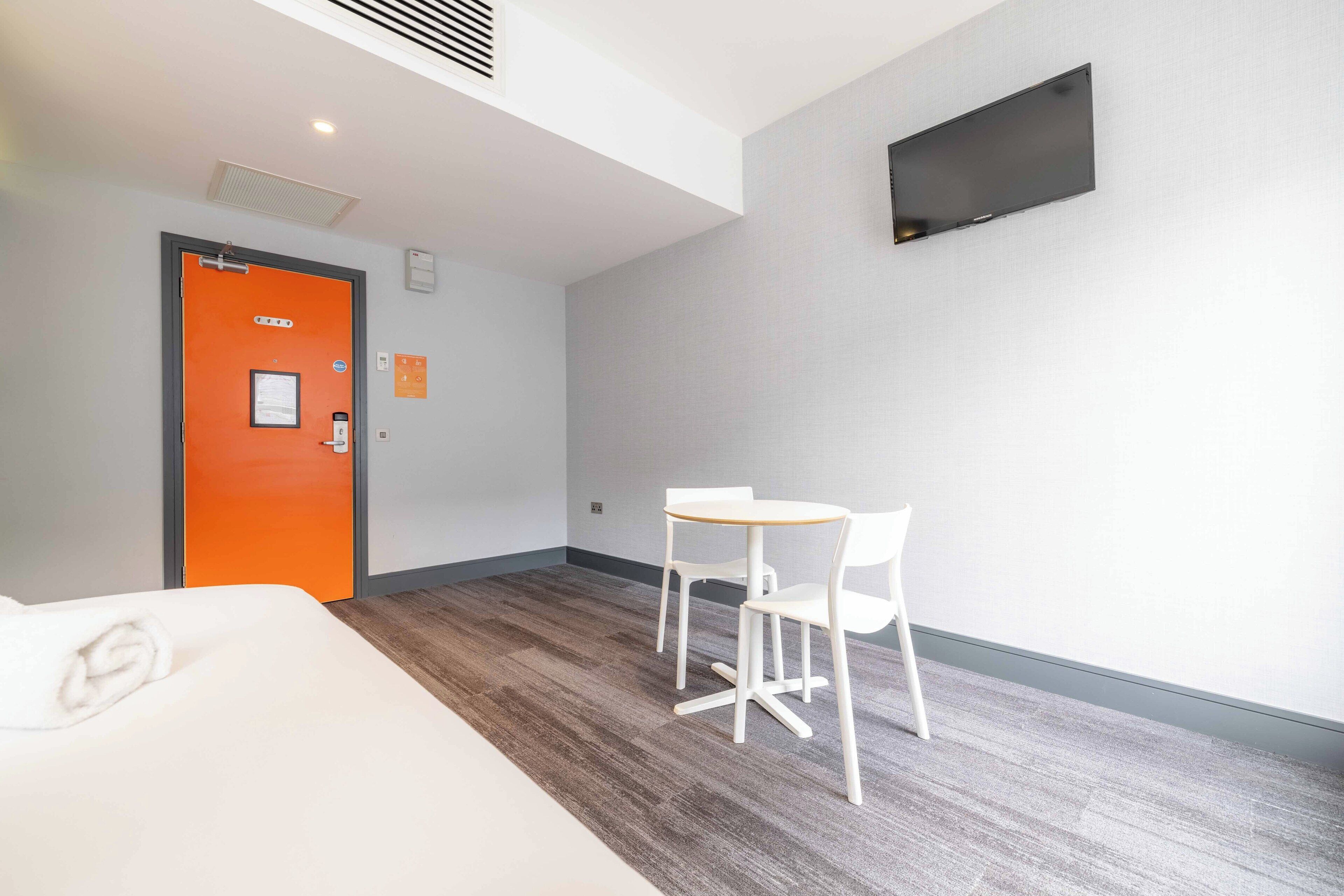 A hotel room with a bed, an orange door, a small table with chairs, and a flat-screen TV mounted on the wall.