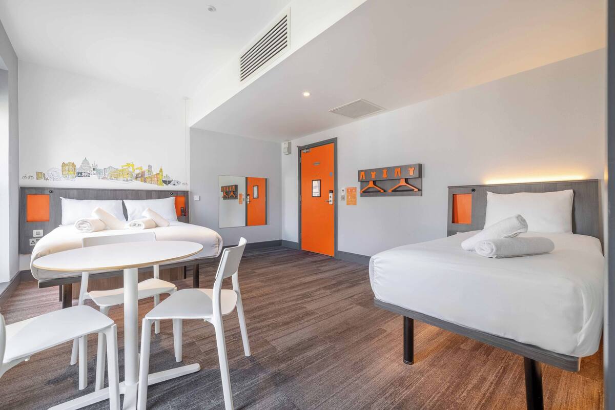 A hotel room with a bed, a desk, and chairs. There is a door with an orange frame and a mirror above it.