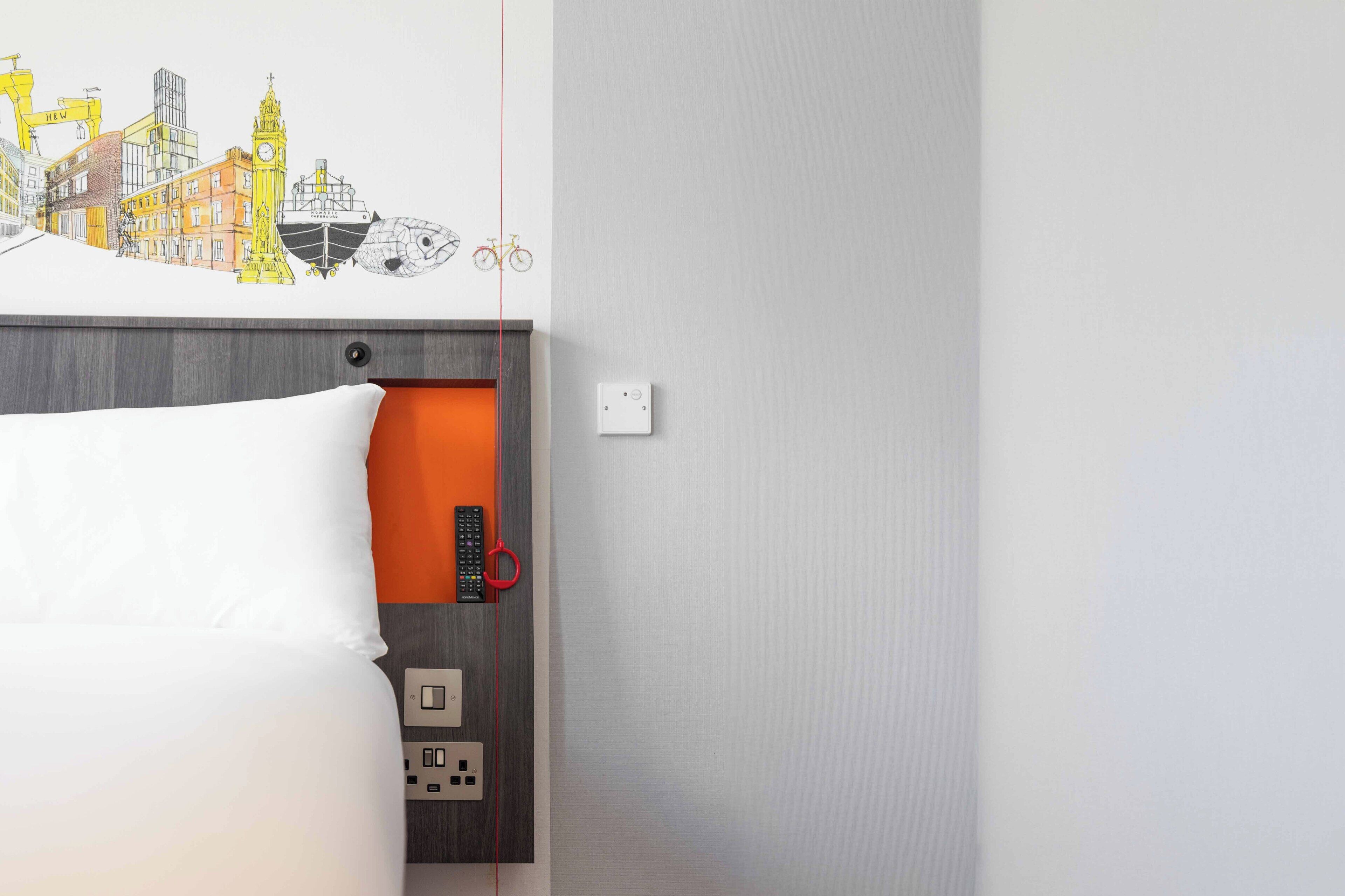 Une chambre d’hôtel avec un lit, une fresque murale représentant un paysage urbain, une télécommande rouge et une prise électrique.