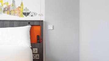 Une chambre d’hôtel avec un lit, une fresque murale représentant un paysage urbain, une télécommande rouge et une prise électrique.