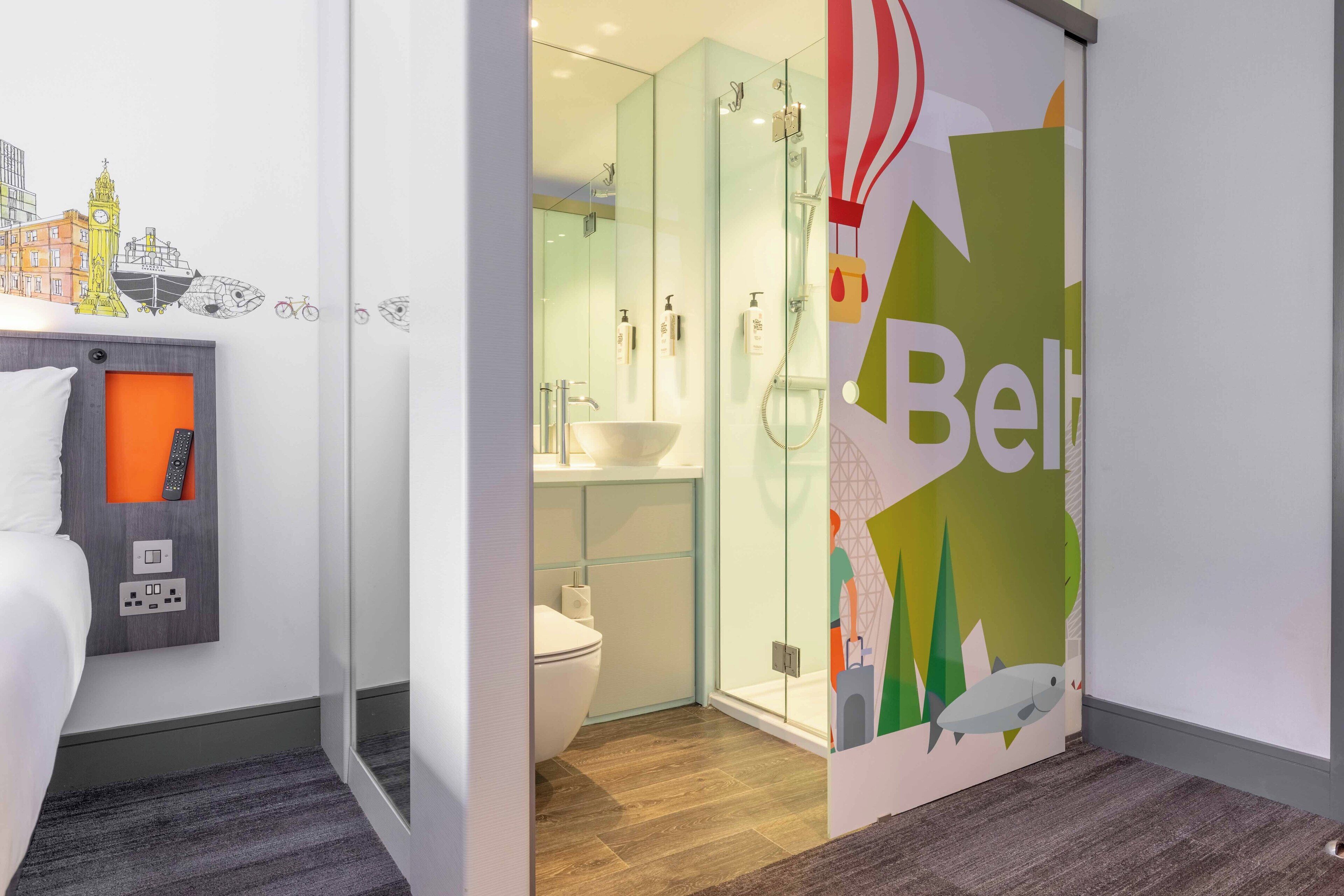 A modern hotel bathroom with a glass shower enclosure, a white sink, and a toilet. There is a colorful wall decal with the word "Belt" and a cityscape illustration.