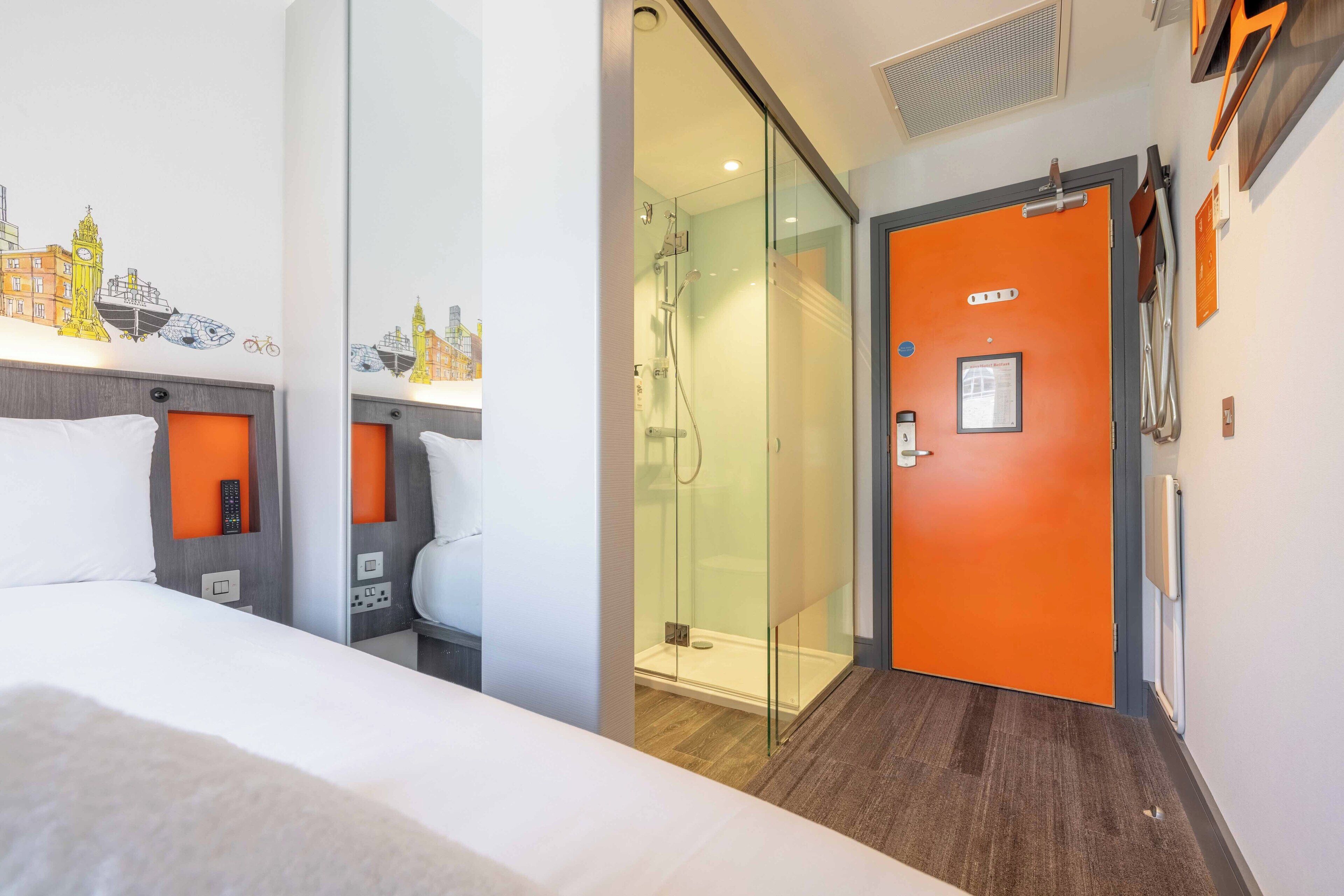 A hotel room with a bed, a shower, and a glass door leading to a bathroom.