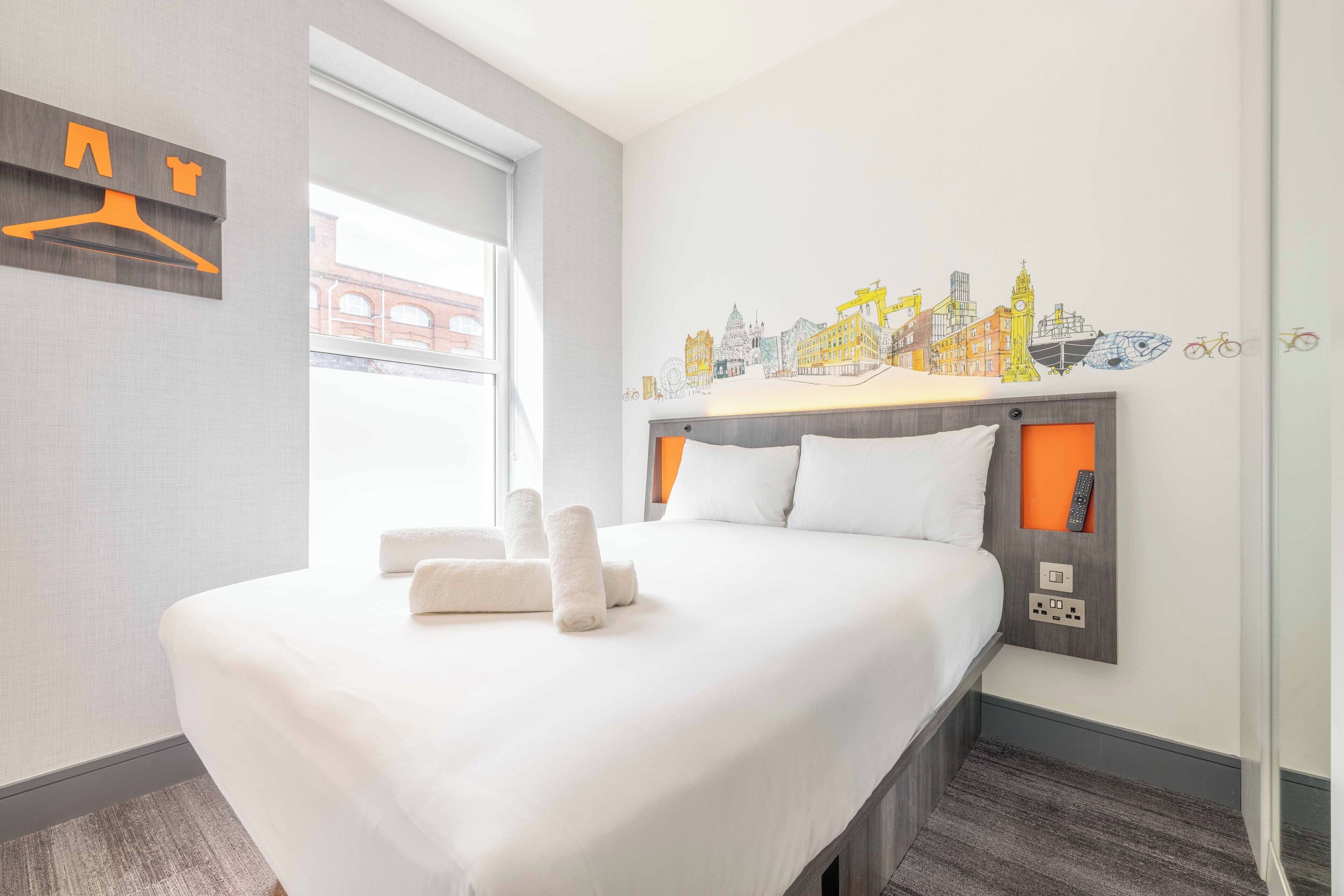 Une chambre d’hôtel avec un lit, du linge de lit blanc, une fresque représentant un paysage urbain, une fenêtre et un interrupteur fixé au mur.