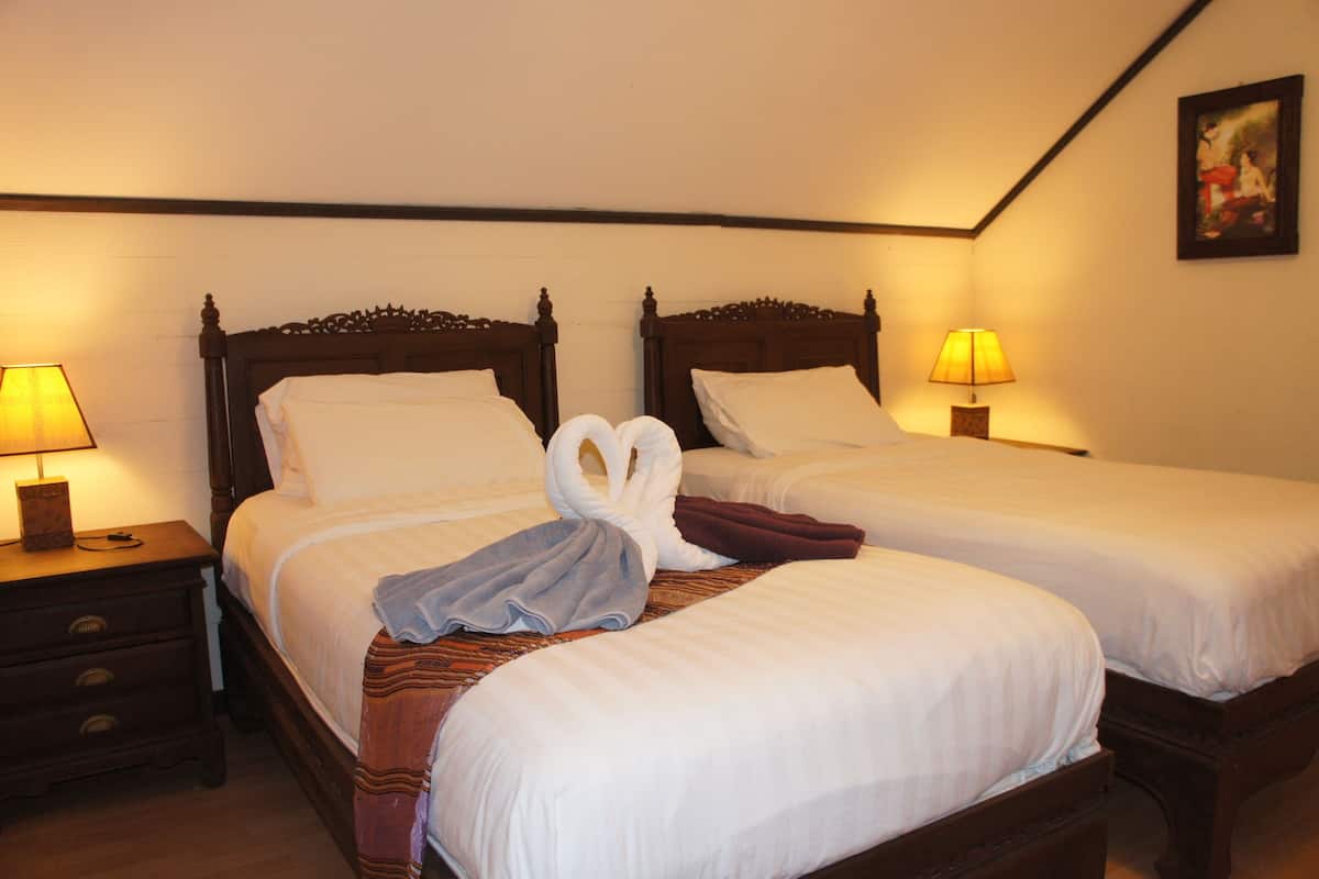 A hotel room with two single beds, each with a wooden headboard, white linens, and a pair of towels arranged in a swan shape on the beds. There are bedside lamps and a nightstand with a drawer.