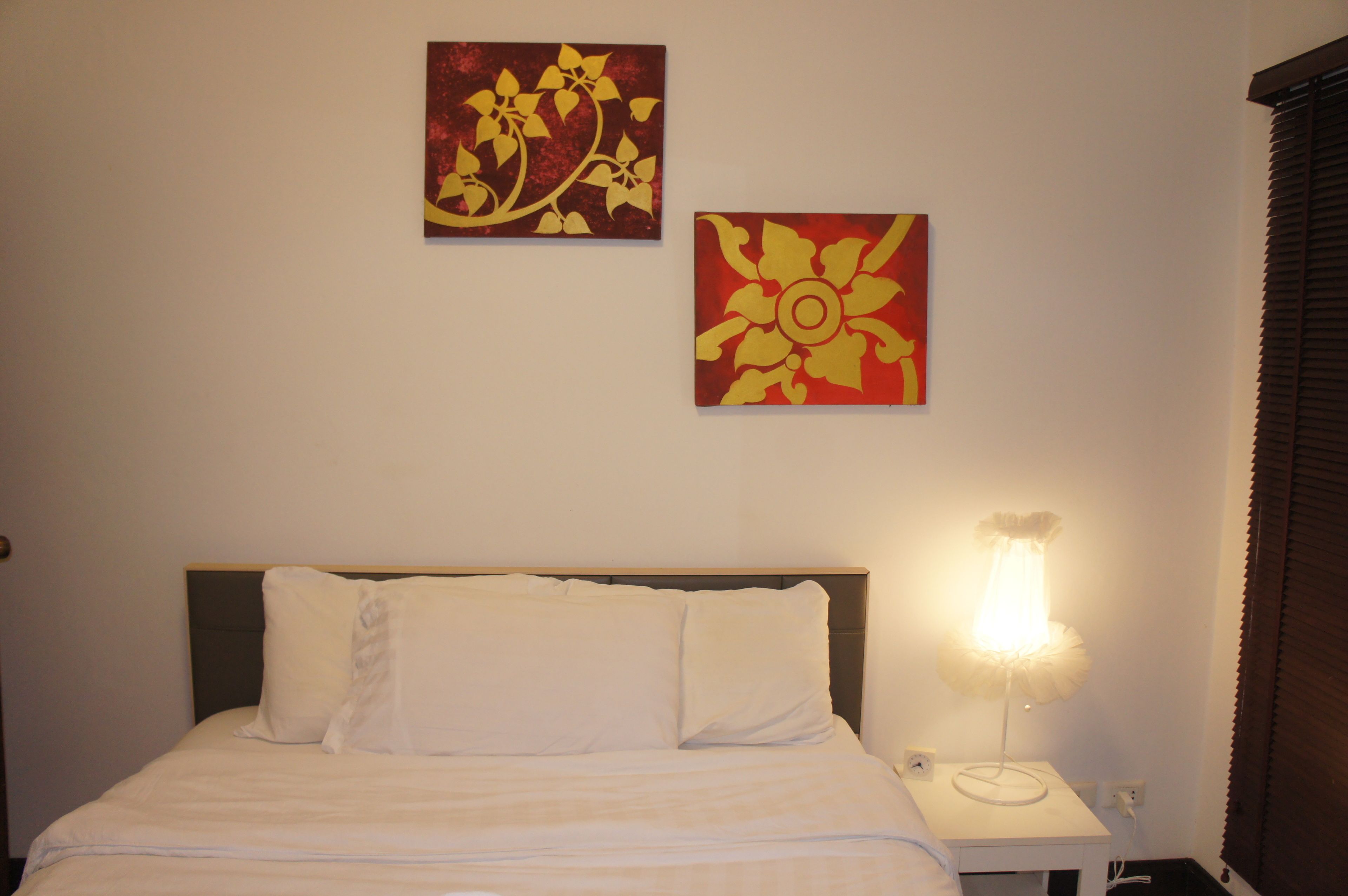 Ein ordentlich bezogenes Bett mit weißen Leinen, ein Nachttisch mit Lampe sowie zwei Blumenbilder an der Wand.