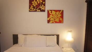 Ein ordentlich bezogenes Bett mit weißen Leinen, ein Nachttisch mit Lampe sowie zwei Blumenbilder an der Wand.