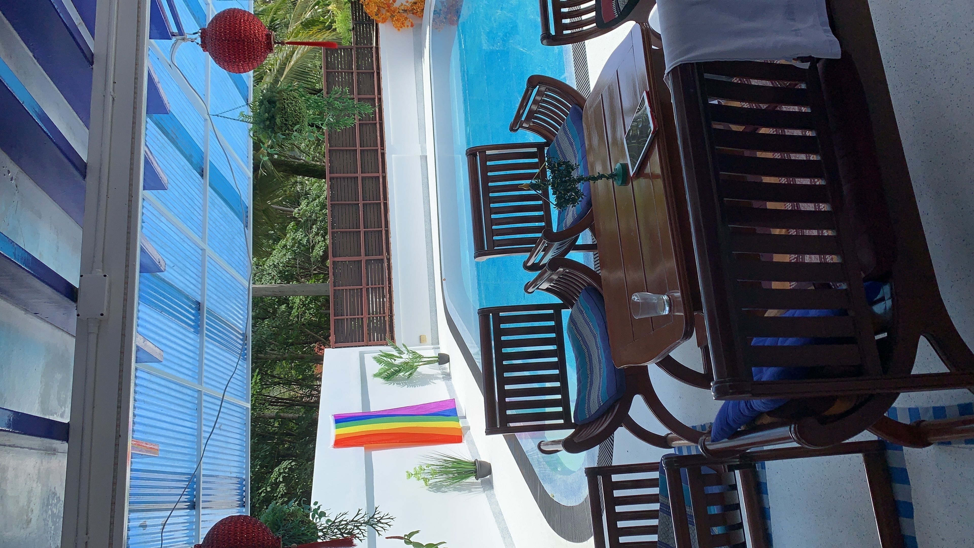 A poolside area with wooden lounge chairs and a table, a blue building with a corrugated metal roof, and a rainbow flag.