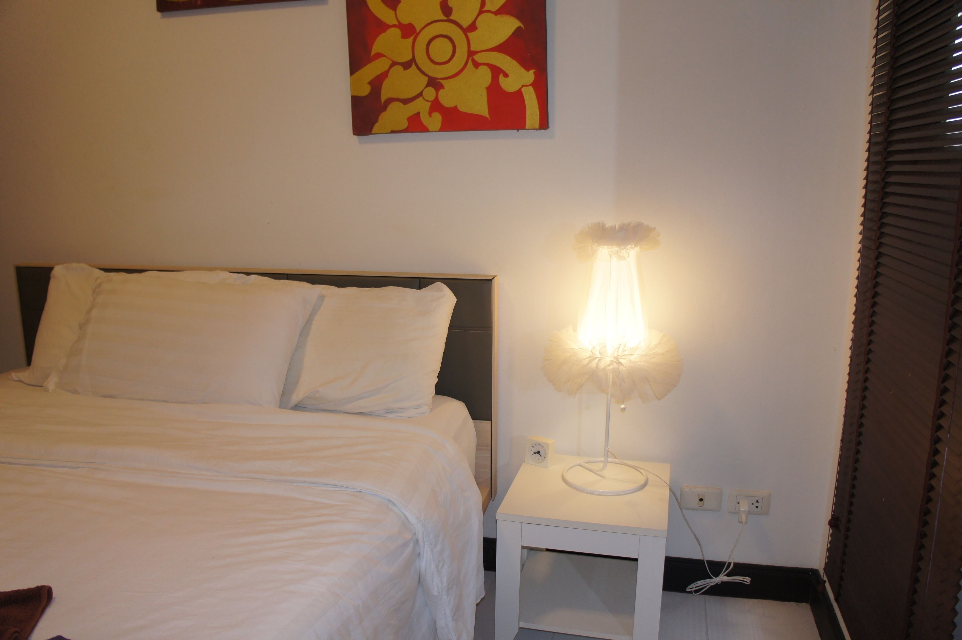 A neatly made bed with white linens, a bedside table with a lamp, and a wall clock.