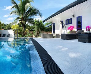 A swimming pool with a black edge, surrounded by outdoor furniture and a white building with blue-framed windows. - Phuket Gay Home Stay - Caters to Men (Kathu)