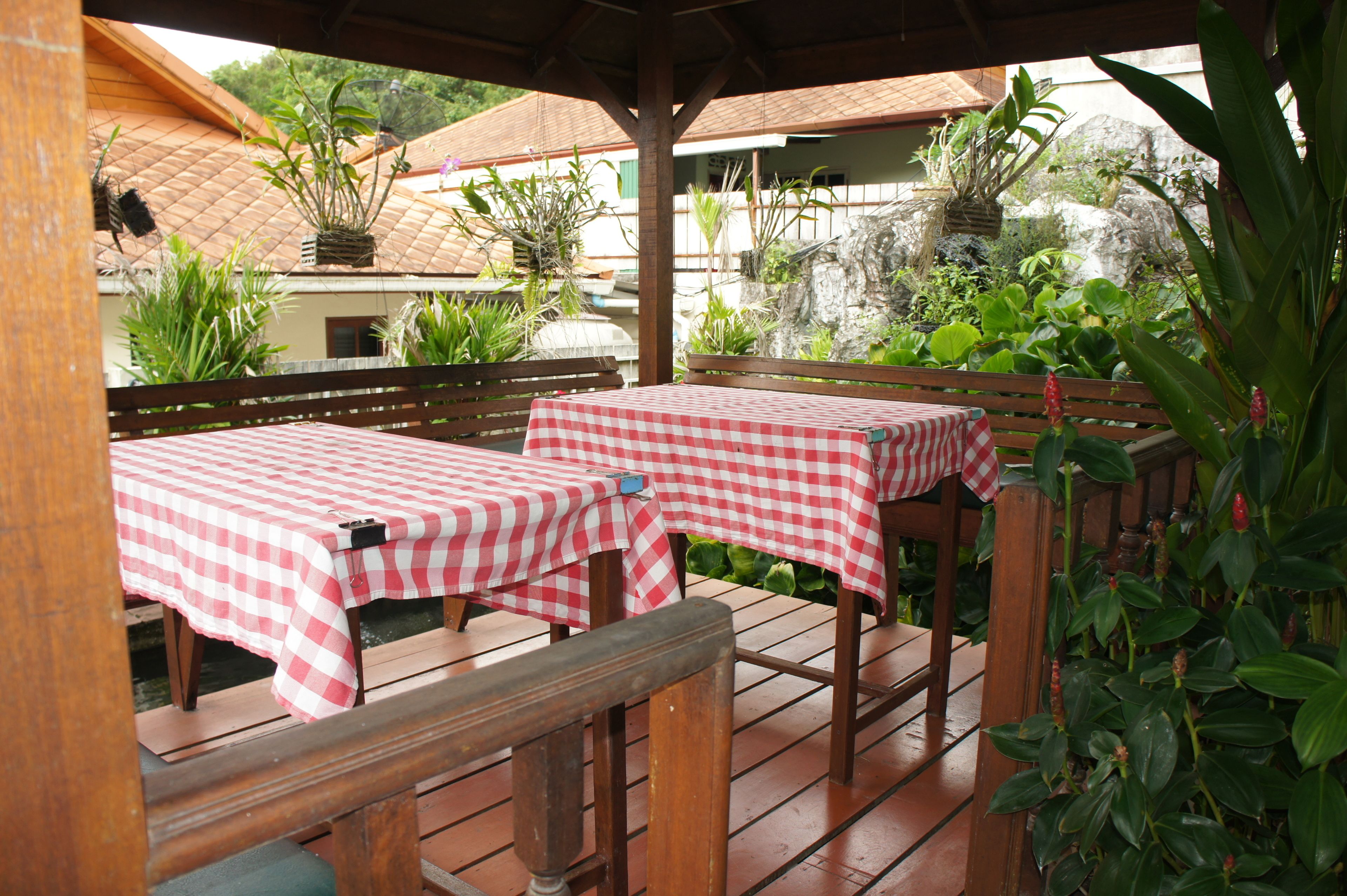 An outdoor patio with two checkered tables and benches, surrounded by lush greenery and a rustic wooden structure.