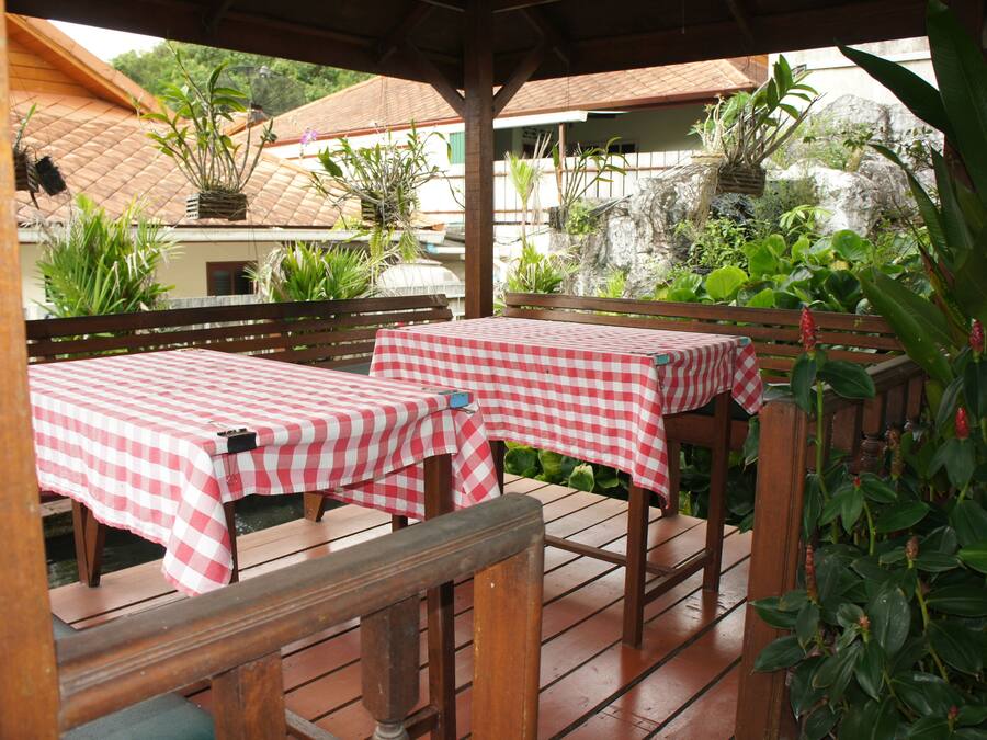 An outdoor patio with two checkered tables and benches, surrounded by lush greenery and a rustic wooden structure.