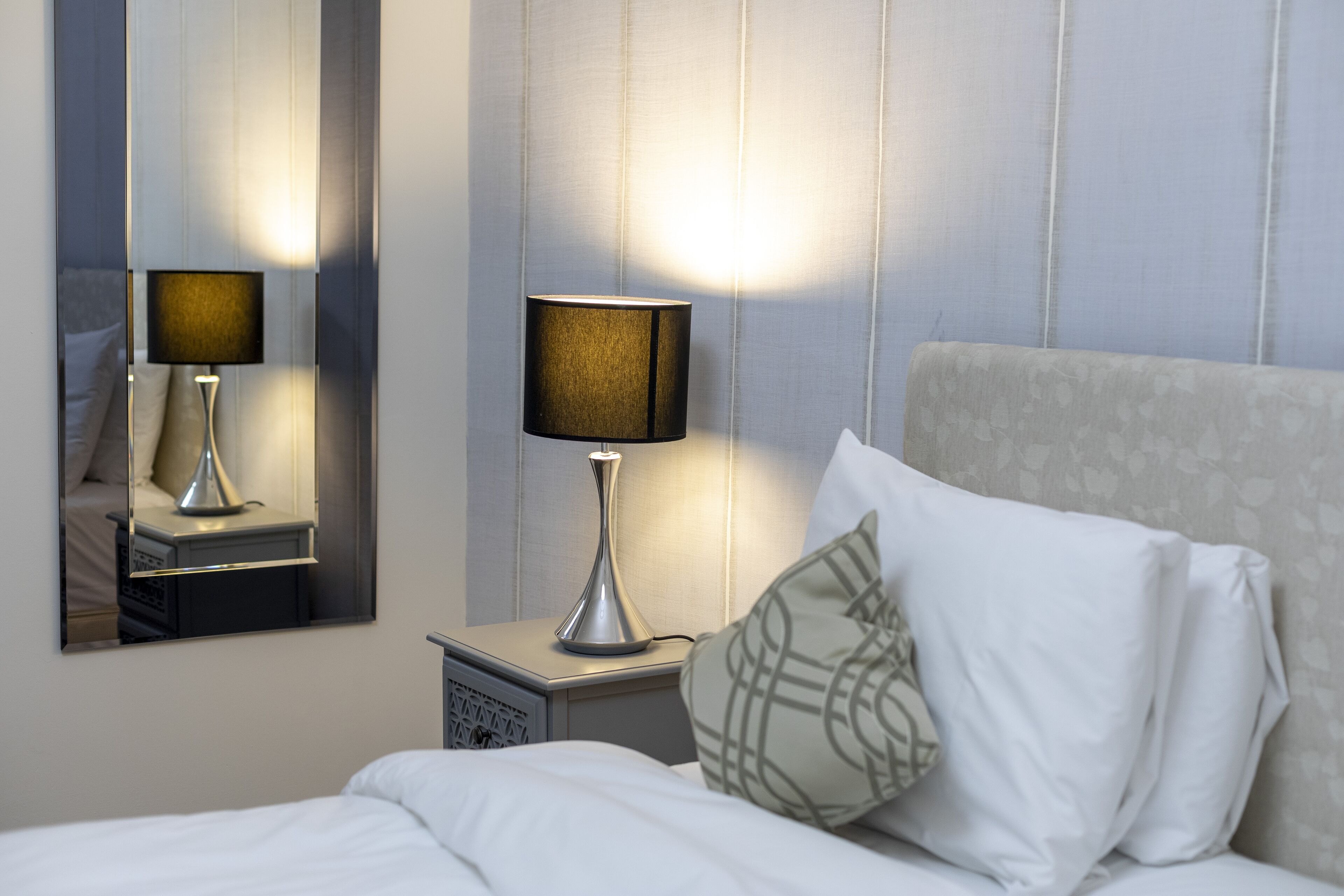 Una cama bien hecha con sábanas blancas, una lámpara de noche y un espejo que refleja la habitación.