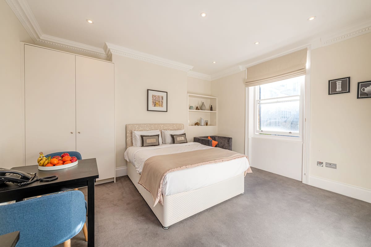 A bedroom with a bed, a desk, a chair, a bowl of fruit, and a window.